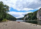 Der Kiesstrand an der Donau ist mit vereinzelten Sonnenanbetern belegt. Rechts fließt die Donau und dahinter ragen die Felswände auf. Der Himmel wäre strahlend blau, wenn nicht die vielen Wolken wären.