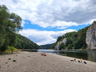 Kiesstrand an einem Fluss mit Menschen, die sich sonnen. Im Hintergrund bewaldete Hügel und Felsen.