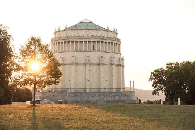 Rundes historisches Gebäude mit Statuen auf dem Sims, Sonne hinter Baum im Park bei Abendlicht