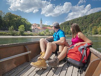 Paar sitzt in einem Boot auf einem Fluss und blickt auf ein Gebäude mit Turm und Wald im Hintergrund.